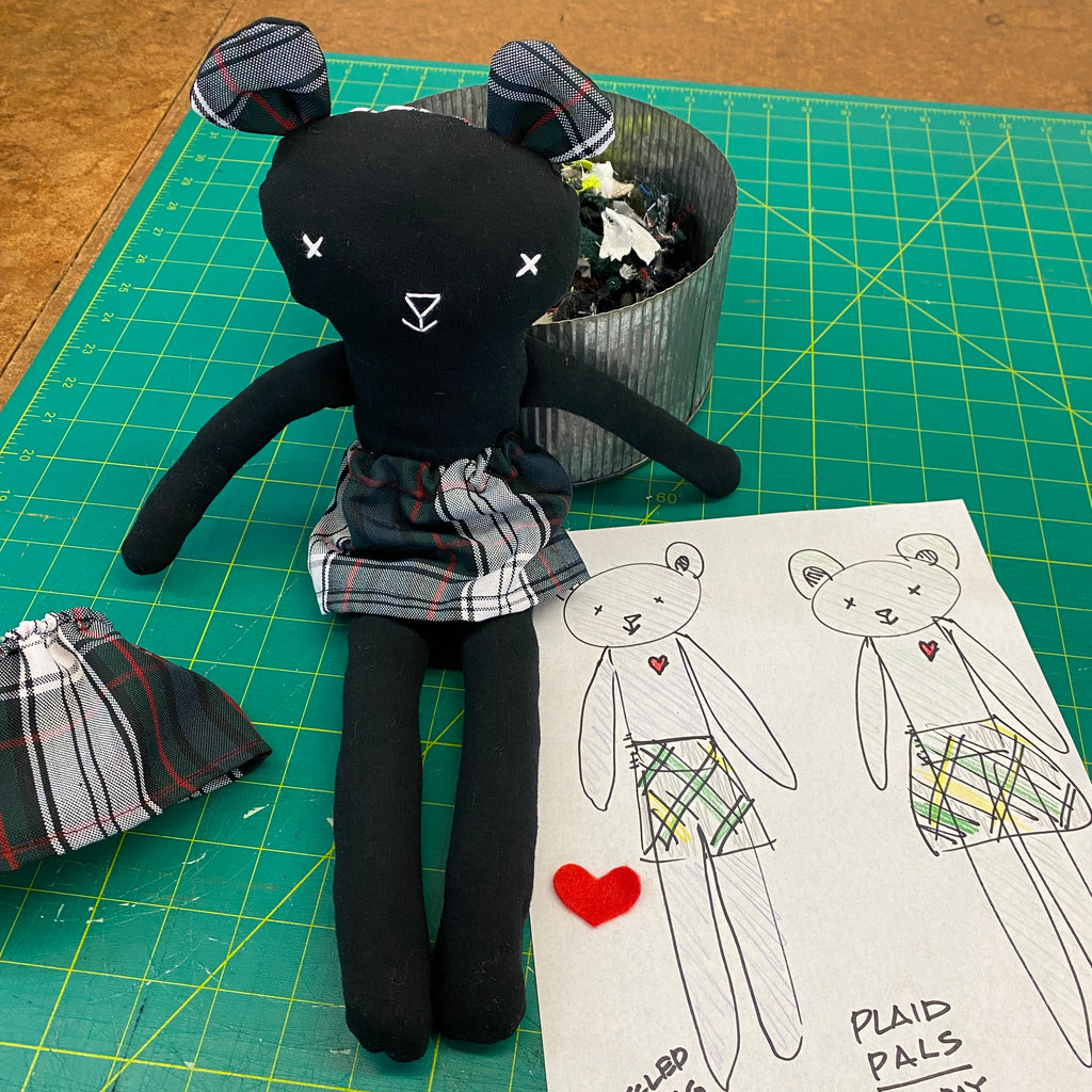 Black plush toy with plaid pants on a green cutting mat with a drawing of a similar toy.